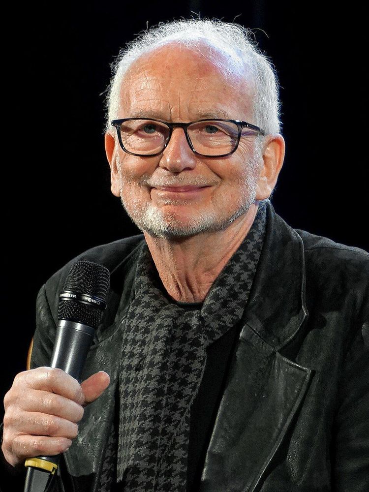 Photo d'Ian McDiarmid, portant des lunettes, tenant un microphone, vêtu d’une veste noire et d’un haut avec un motif hachuré. Le fond est sombre.