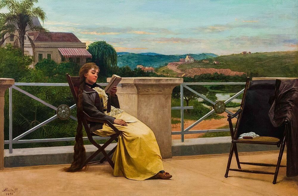 Une peinture à l'huile intitulée "La Lecture" par José Ferraz de Almeida Júnior, datant de 1892. L'œuvre représente une femme assise sur une chaise en bois sur une terrasse, en train de lire un livre. Elle porte une robe longue et jaune, un chemisier clair et une veste foncée. Ses longs cheveux bruns sont visibles derrière elle. La terrasse est bordée d'une balustrade blanche et donne sur un paysage verdoyant avec des collines, des maisons, et ce qui semble être un plan d'eau ou une rivière au loin. Un palmier est visible sur le côté gauche de l'image, et une autre chaise pliante sombre est placée à droite, avec un vêtement drapé dessus. Le ciel est nuageux avec des teintes de bleu et d'orange pâle.