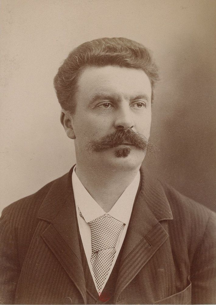 Portrait de Guy de Maupassant, photographié en noir et blanc, vêtu d’un costume formel avec une cravate. Le fond est uni et clair, mettant en avant le sujet principal.