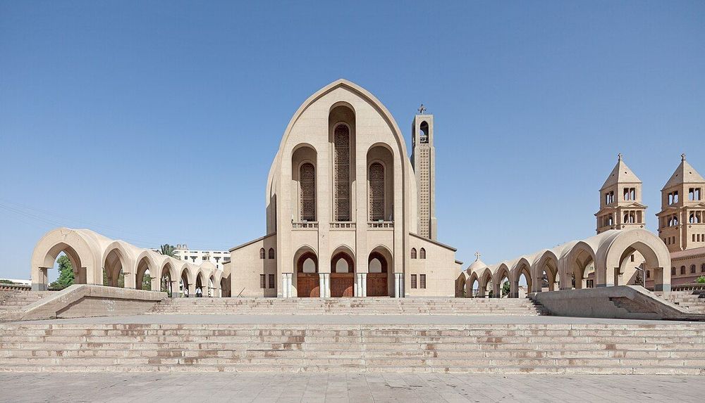 Grande image frontale de la cathédrale copte orthodoxe Saint-Marc du Caire. La cathédrale est un grand bâtiment moderne en pierre de couleur sable avec une grande arche en ogive en son centre. Deux portes d'entrée en bois, en partie à l'ombre, sont visibles sous l'arche principale. Deux longues fenêtres cintrées et grillagées se trouvent au-dessus. De chaque côté, un long couloir avec des arcs de cercle en plein cintre mène au bâtiment principal. Une rangée d'escaliers en pierre mène à la cathédrale. Le ciel est bleu et clair.