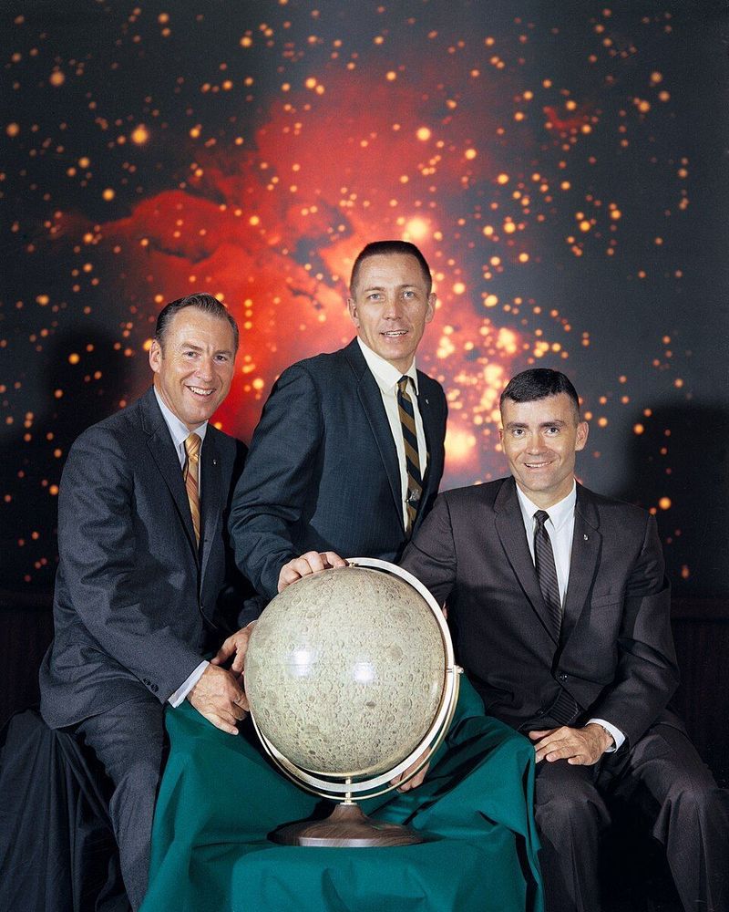 Photographie de trois membres de l’équipage d’Apollo 13. Ils posent devant un fond spatial éclatant, vêtus de costumes formels et tiennent ensemble un globe lunaire sur une table recouverte d’un tissu vert. Le fond représente une image spatiale avec des étoiles et des nébuleuses brillantes dans diverses teintes d’orange et de rouge.
