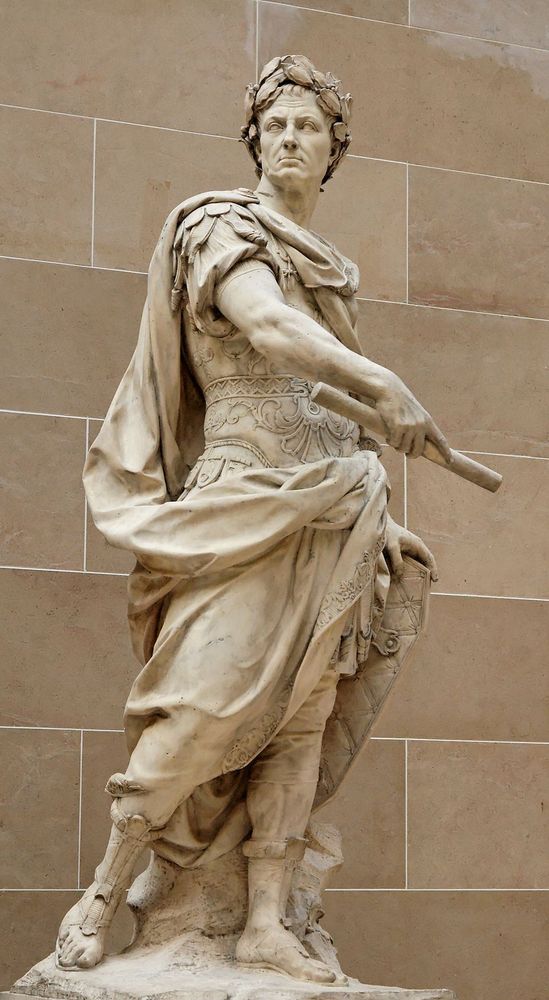 Statue en marbre de Jules César réalisée par Nicolas Coustou en 1713. La sculpture représente César debout, vêtu d'une tenue romaine traditionnelle, avec une cape drapée sur son épaule gauche. Il tient un rouleau dans sa main droite, symbolisant un document ou un décret. Les détails minutieux, comme les plis de la cape et les ornements de la ceinture, mettent en valeur l'élégance et le savoir-faire artistique de l'époque.