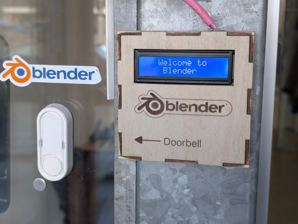 Photo of a laser-cut wooden box. It has an LCD display reading "Welcome to Blender". Engraved are the Blender logo and an arrow pointing to the doorbell.