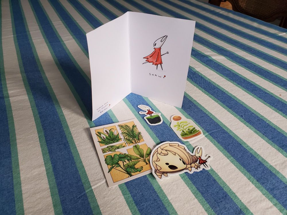 A collection of small stickers, mostly featuring Hornet from Silksong and also plant life, arranged in front of an open greeting card with Hornet, captioned "Shaw!", on a teal and white-striped tablecloth