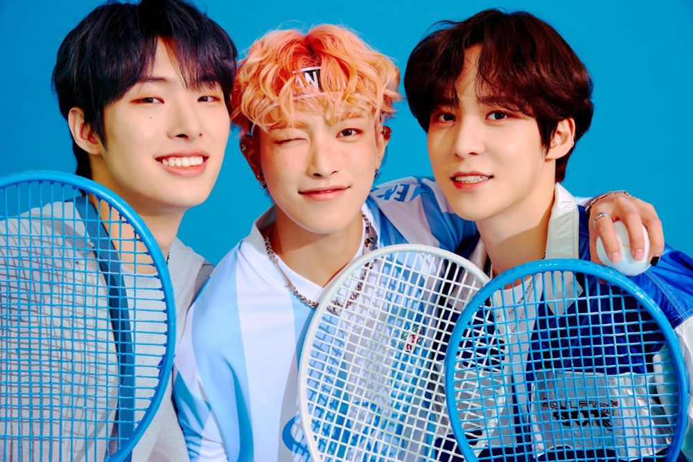 Hongjoong in between Mingi and Yunho. His hair, styled to look curly, is a peachy orange, darker at the roots than the ends.