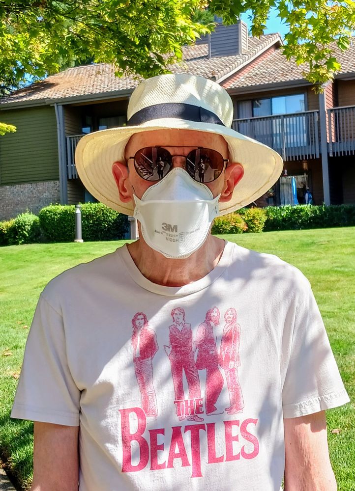 Michael Collins Morton standing in the sunshine, wearing a sun hat, dark glasses, a respirator mask, and a tan shirt with a red picture of The Beatles on the front.