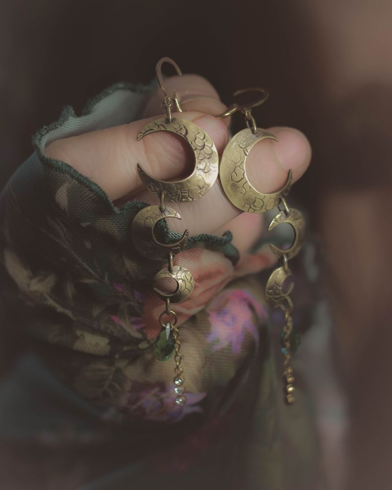 Boucles d'oreilles artisanales en laiton. Les boucles se composent de 3 croissants de lune du plus grand au plus petit, chacun dans un sens opposé au précédent. 
La boucle se termine par un cristal de vert de couleur verte et par j e petite chaîne avec au bout une breloque scintillante de trois brillant blancs. 
La paire est présentée dans une ambiance sombre et lumineuse à la fois portée.