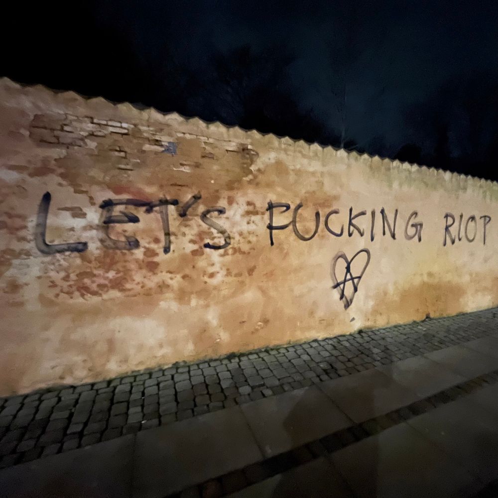 Image of a yellow wall at night where someone has written “let’s fucking riot” signed with a circle-a in a heart