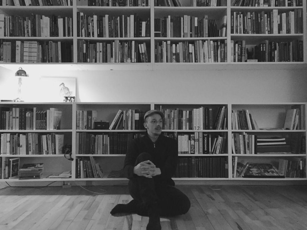 A black and white photograph showing a person wearing a hat sitting on the floor in front of a book case.
