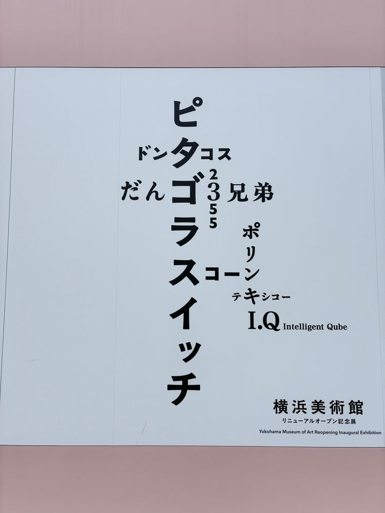 横浜美術館リニューアル記念展「佐藤雅彦展」の掲示。中愛に縦書きで「ピタゴラスイッチ」と書かれているところに、同じ文字が重なる形で「ドンタコス」「だんご3兄弟」「スコーン」などのテキストが横書きになっている。