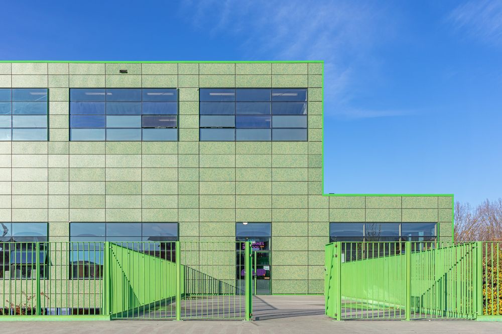 A photo of a school building taken on a sunny day with a blue sky above. The building is a striking, light green colour, with rectangular facade panels covering the entirety of its walls. Bright green railings are visible in the foreground leading to one of the entrances.