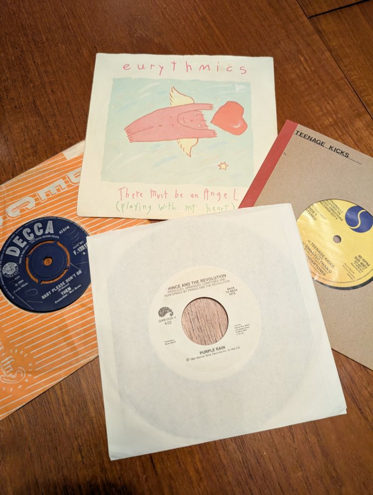 Four seven inch vinyl singles on a wooden table. They are: There must be an angel by Eurythmics, Baby please don't go by Them, People Rain by Prince and the revolution, and Teenage kicks by The Undertones.