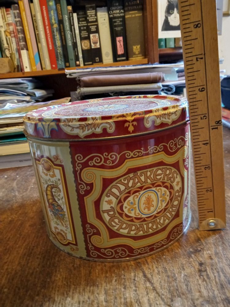 A richly decorated biscuit tin labelled "Nyakers Pepparkakor". A ruler next to it shows that it is just under six inches tall.