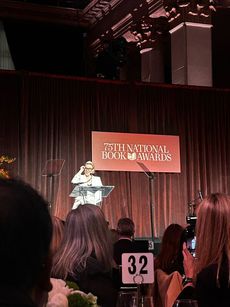 Comedian Kate McKinnon standing on the stage at the National Book Awards putting on her glasses which she has already told the audience are not real and just to make her look smart. 