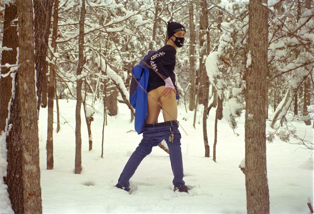 A Femboy standing partially clothed in a snowy forest while spreading his ass cheeks