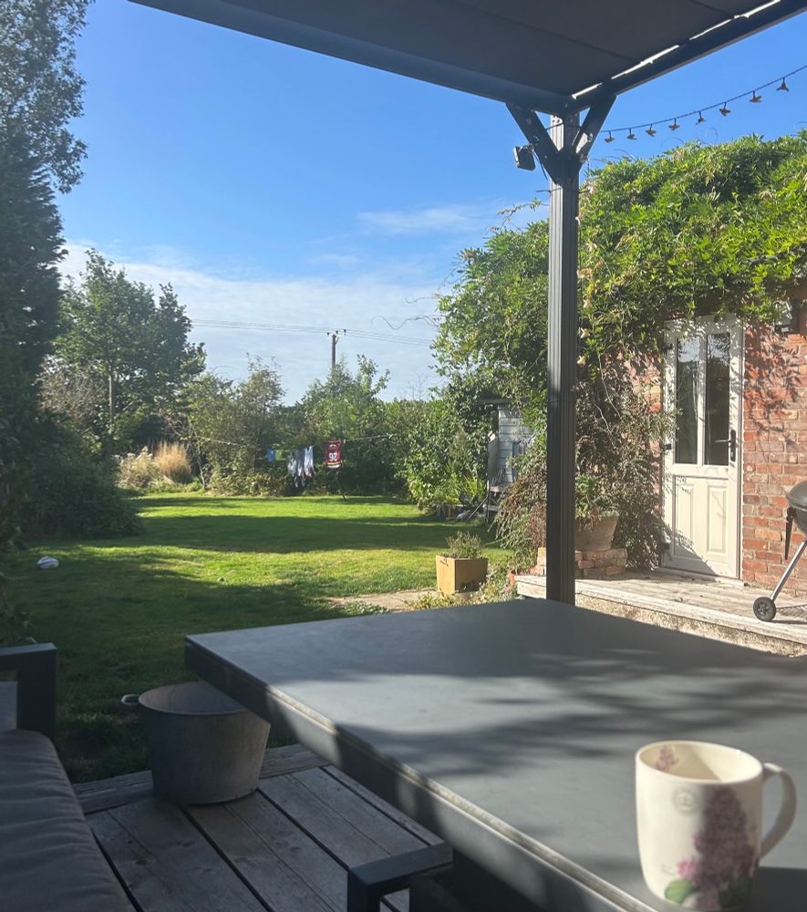 View of the sunny garden from the outdoor sofa under a gazebo. A cup of tea in the foreground on a grey table. Sunshine and shadows play across the lawn, shrubs, wild long grass and thistle down and trees. A few clothes hang on the overspill washing line. 