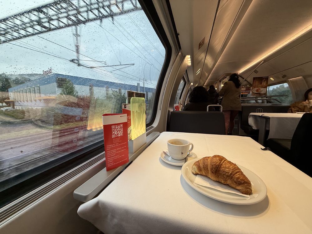 Kaffee und Croissant auf einem Tisch im Speisewagen, links das Fenster, aus dem Gebäude zu sehen sind. Bewölkter Himmel ist zu erkennen.