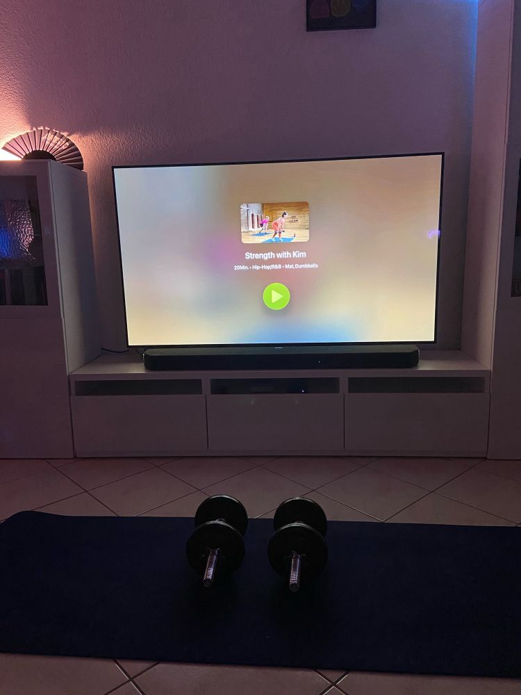 Ein Paar Einhandhantel auf einer Sportmatte. Im Hintergrund ein TV Gerät mit Apple Fitness+ Programm