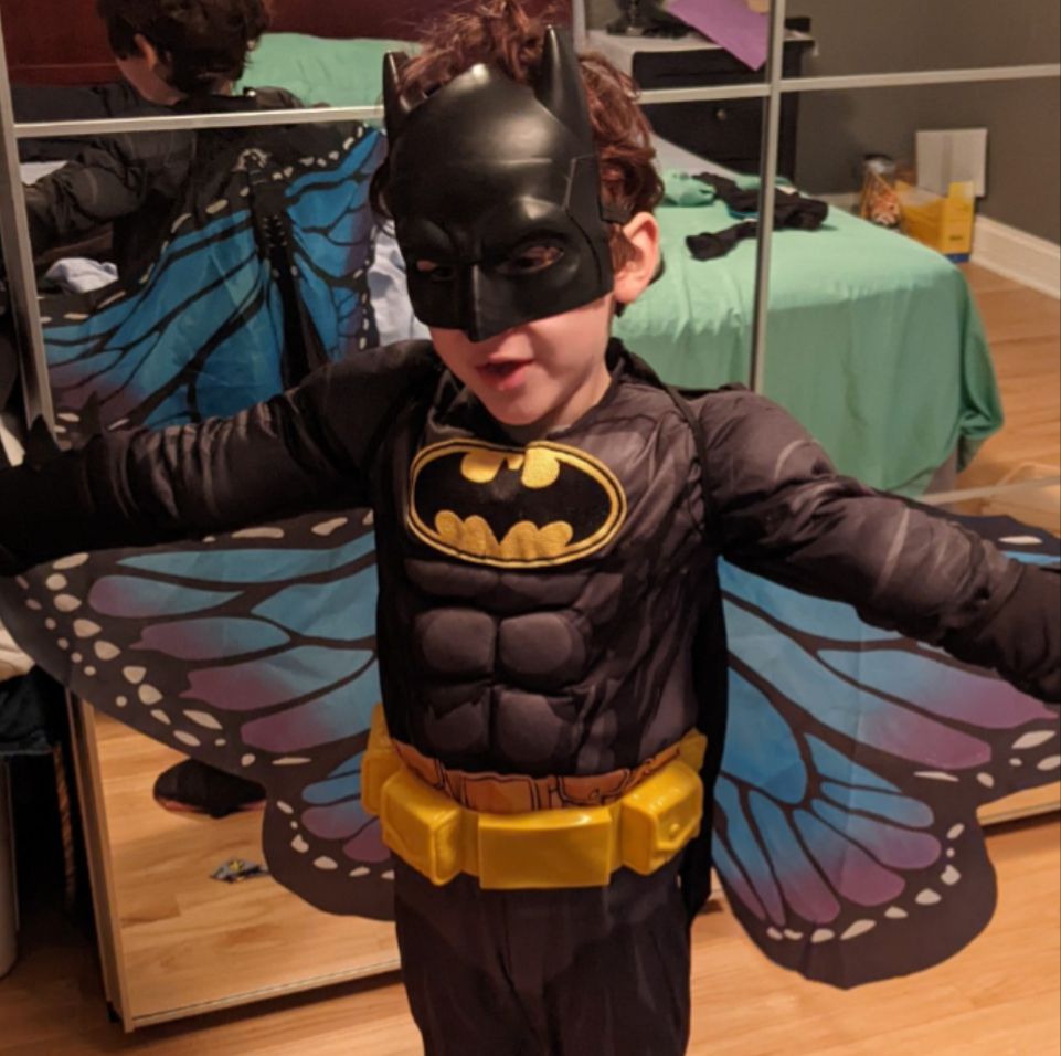 Child dressed in a Batman costume with a mask, yellow belt, as well as some pretty blue and purple flowy butterfly wings. Extremely messy bedroom unfortunately reflected in the mirror behind.