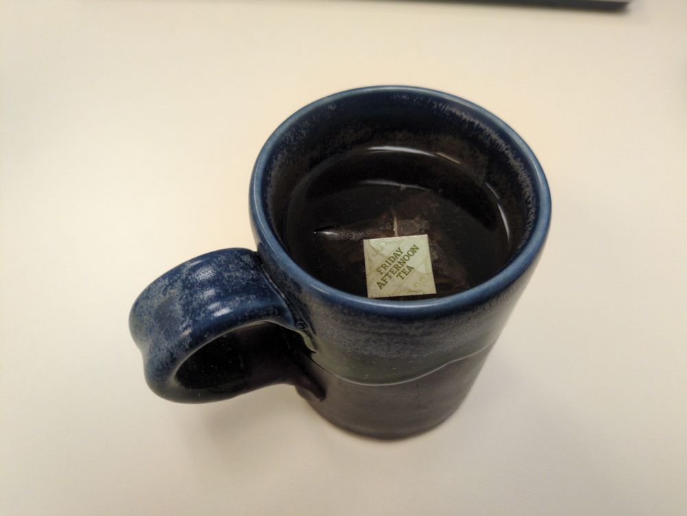 Tea bag from Friday Afternoon Tea (unsmoked lapsang) floating in what could be described by some as tea. Fourth time this bag has been re-steeped. Cup is glazed blue and brown against a depressing white background. 