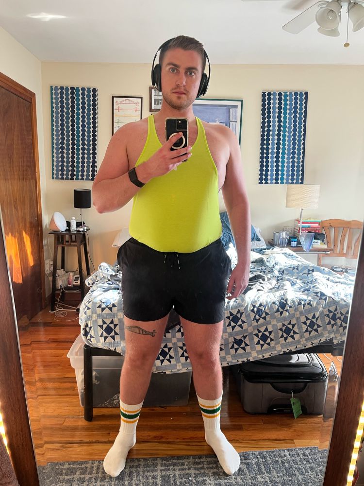 Sam in neon green stringer tank, short black shorts, and green and gold striped crew socks in his bedroom mirror after going to the gym