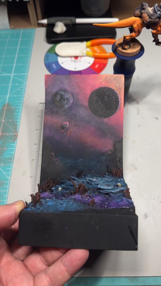 A miniature painting diorama of a pink and purple sky with three planets. The foreground is an Allen landscape with shades of blue and a purple stream bisecting it. 