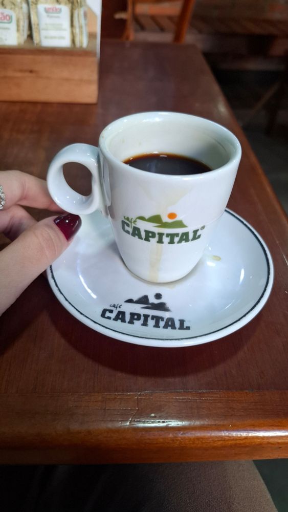 uma xícara de café pequena branca com a logo do café capital do Rio de janeiro (é o pão de açúcar como logo) e minha mão está do lado 