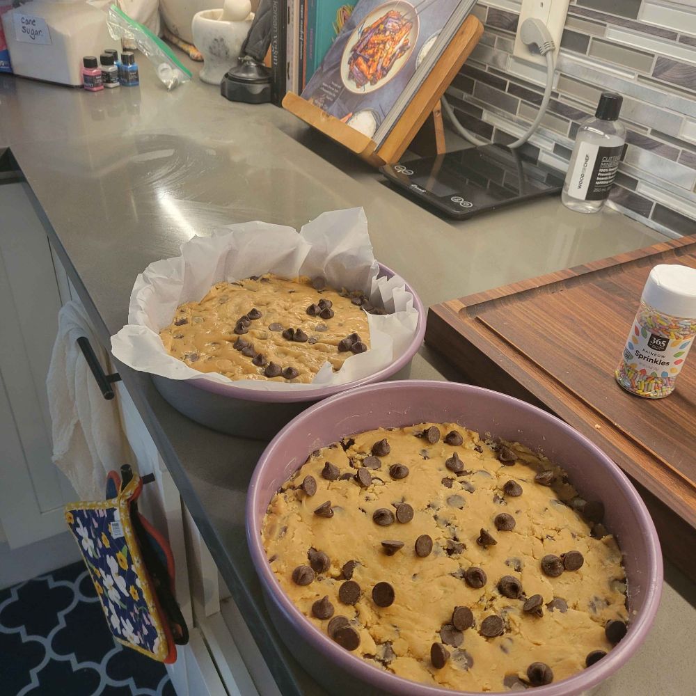 photograph of two round cake tins holding chocolate chip dough for cookie cakes on top of a kitchen counter