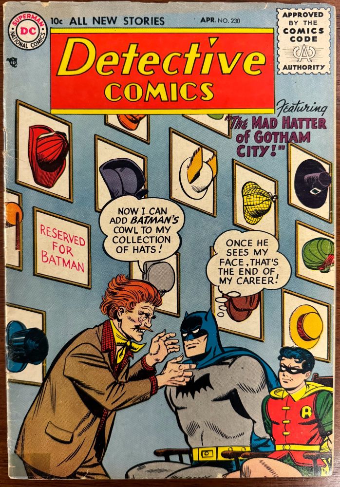 Jervis Tetch, the Mad Hatter of Gotham, has Batman and Robin tied to chairs. He’s about to remove Batman’s cowl to add to his large hat collection displayed on the wall behind him. 