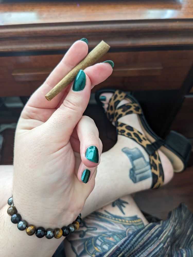 Professor McGovern's pale hands holding a raw paper wrapped blunt betwixt deeply emerald lacquered fingernails. A bracelet of tigers eye encircles their wrist. In the background you can see their tattooed legs and feet in a pair of cheetah print high heels. 

🍃 🧡🖤 🖤🧡 💚