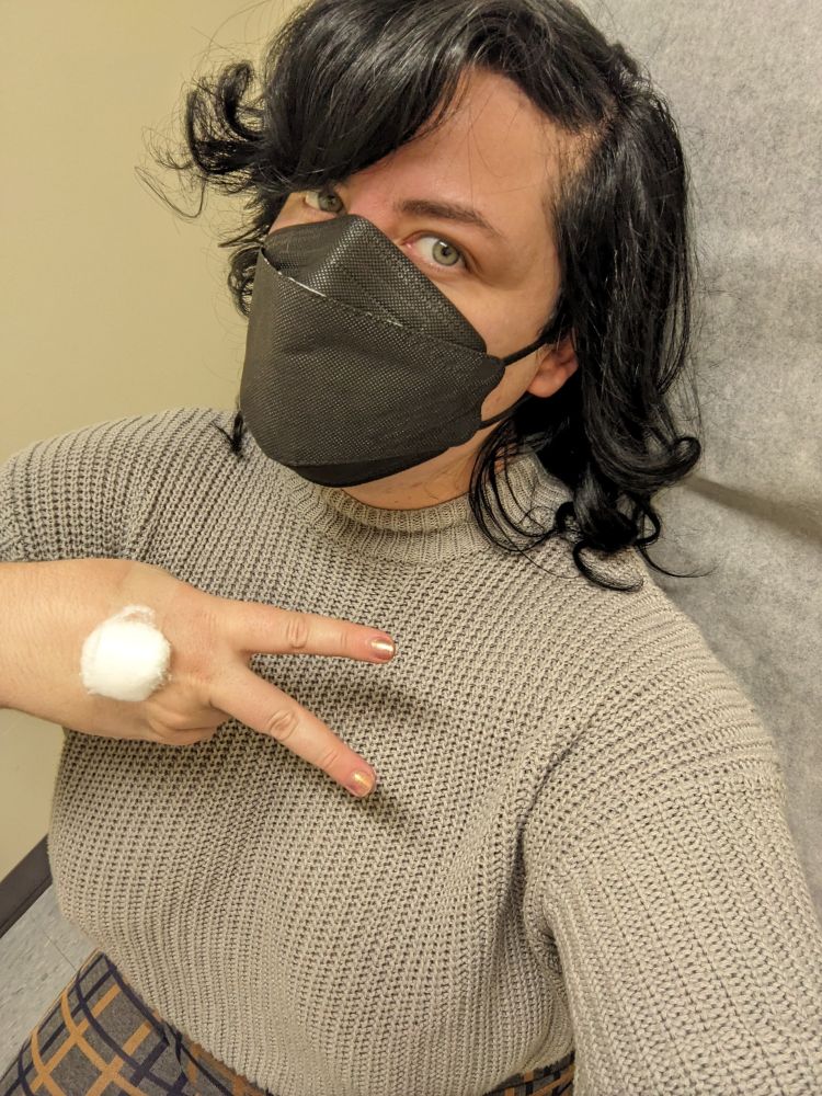 Gwynne with messy curly black hair in a black kn95 mask, showing the back of their hand where there is a taped cotton ball - the site where blood draw for an STD test has been taken. Their green eyes are bright and sparkling.