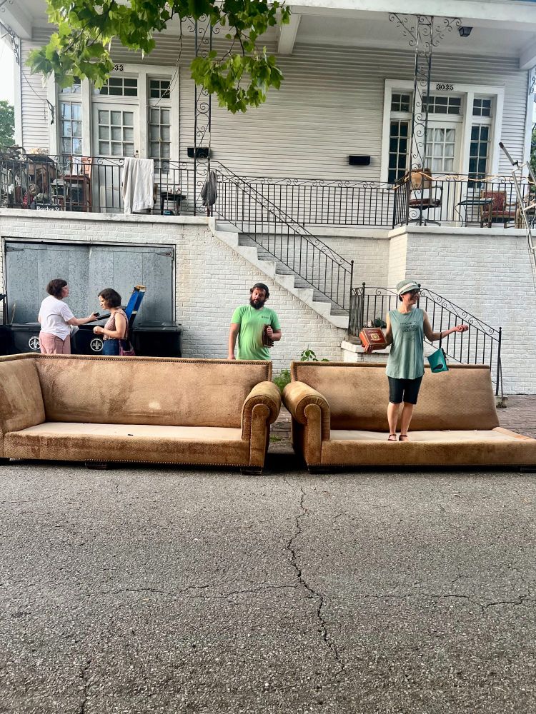 A dirty sectional sofa on the street in front of a white house. Two people behind the sofa are engaged in a conversation about things in nearby garbage cans. Someone in a green shirt is standing behind the sofa and looking at the camera. Another person is standing on one part of the sectional, arms outstretched to the side, holding various items. New Orleans-strength cocktails may have been involved.