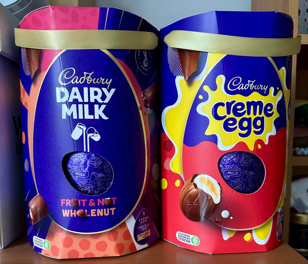 Two Cadbury’s easter eggs. One is in a purple coloured Dairy Milk box. The other is in a purple, yellow and red coloured creme egg box. 
