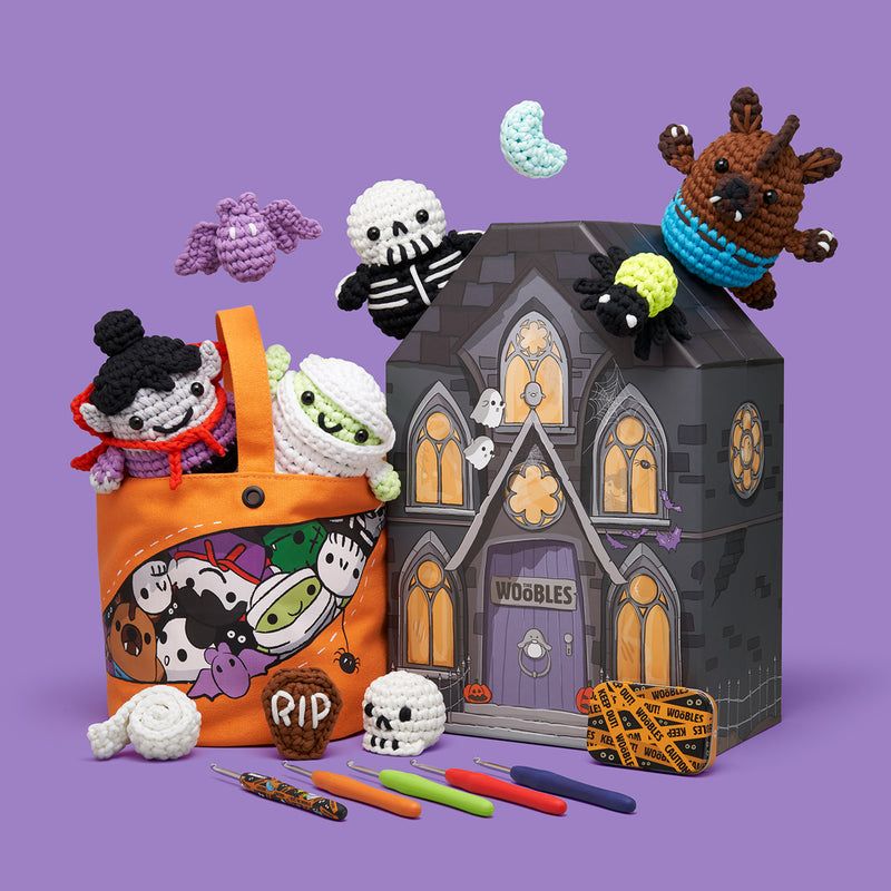 A group of cute, crocheted Halloween characters and creatures are displayed with an orange tote bag, beginner crochet kits, and a haunted house box labeled WOOBLES, all set against a purple background.