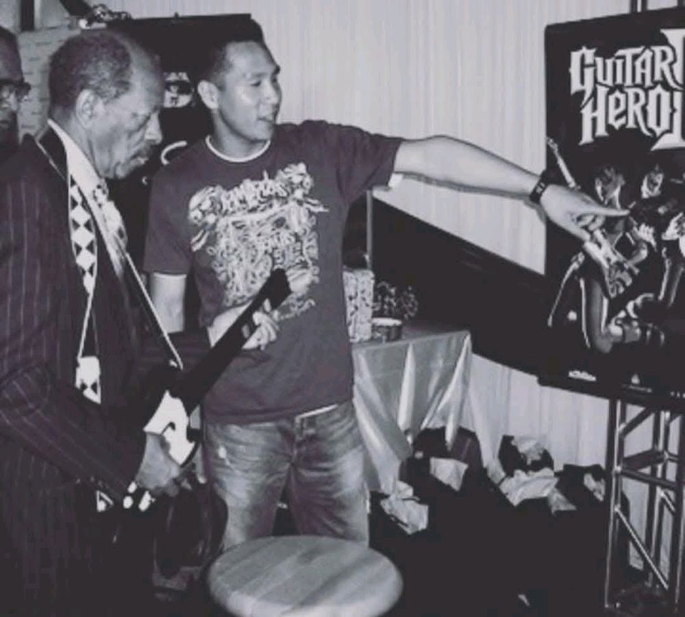 Ornette Coleman playing Guitar Hero, a look of concentration on his face. A younger friend is pointing at the screen as if showing him something 