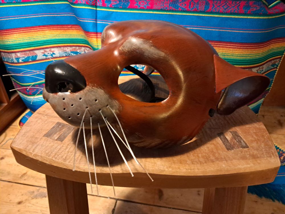 A half mask in the shape of an otter face, painted in chestnut brown with lighter shades around the muzzle, brows and inside the ears. The nose is black and there are nylon filament whiskers. 