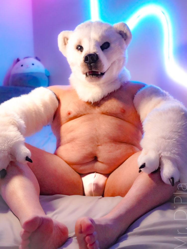 A polar bear fursuiter sits in their underwear on a bed with their legs encircled, waiting for someone to sit in their lap.
