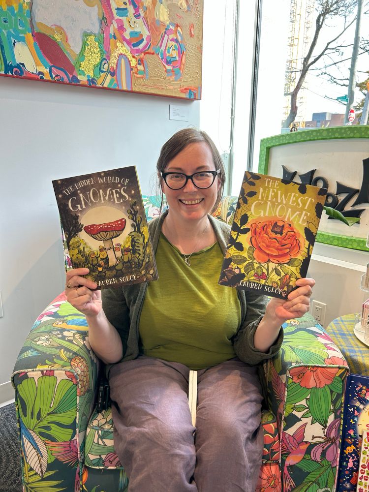 Smiling author holding up their signed books - The Hidden World of Gnomes and The Newest Gnome - in a vibrant floral fabric chair.