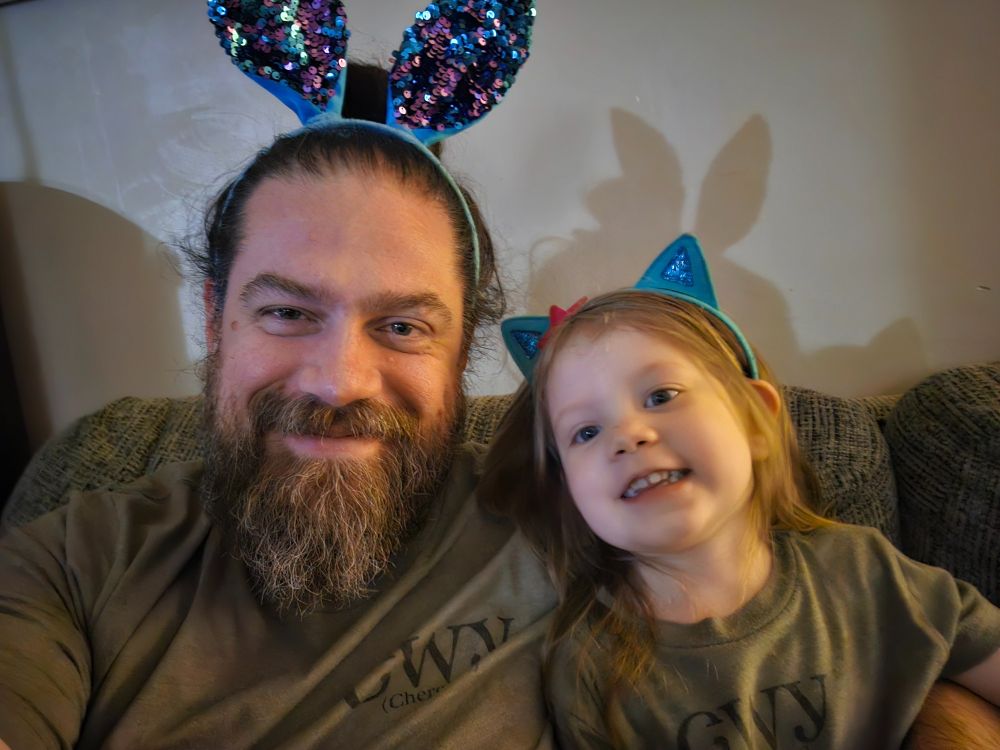 A man wearing a bunny-ear headband poses with his daughter, who is wearing a cat-ear headband. They are wearing matching, green shirts.