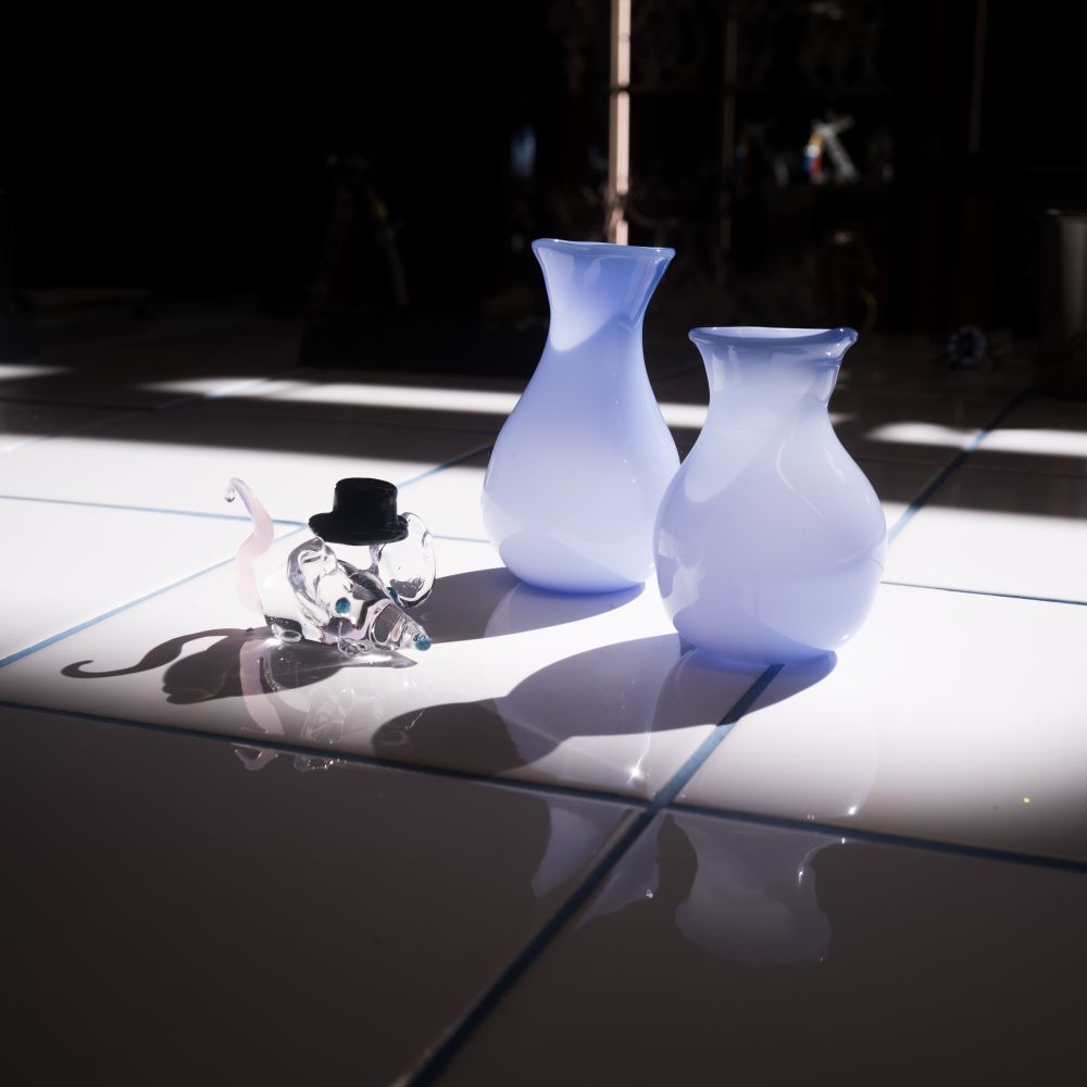 A clear glass mouse with a pink tail and black top hat next to two faded blue vases