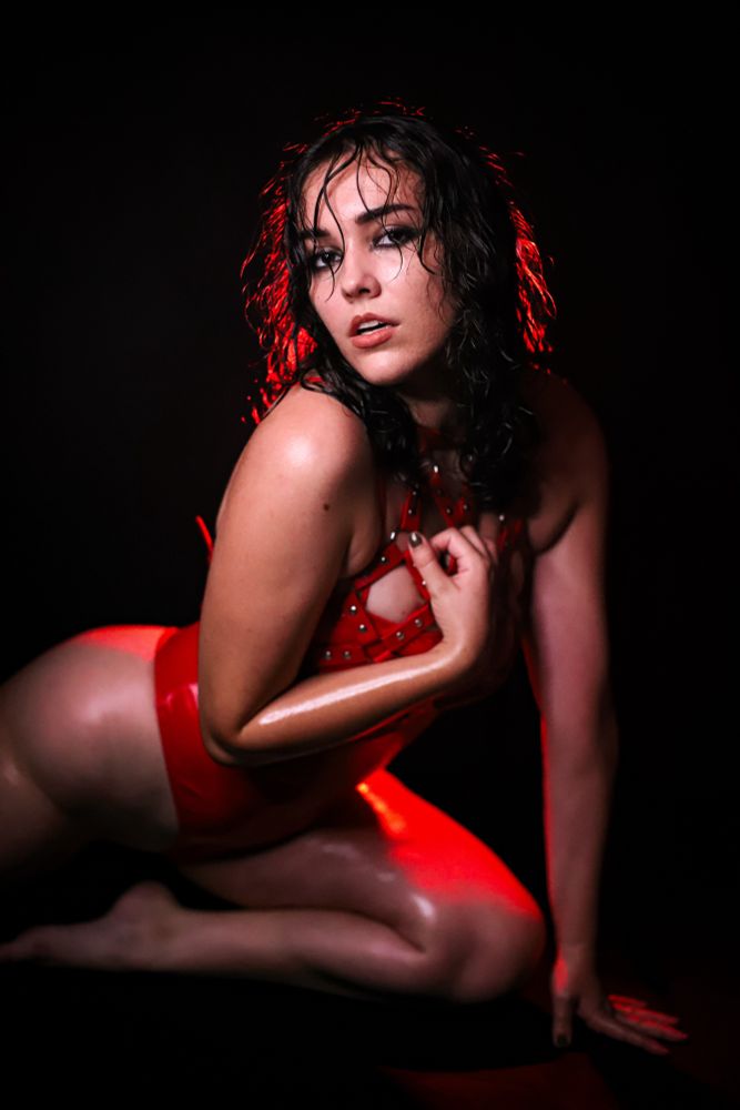 Hailie is wearing a red PVC bodysuit. They appear to be wet, with their skin oiled and hair wet. They are posed on their knees facing the camera.