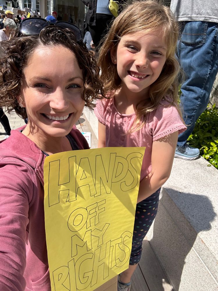 Author Meghan Joyce Tozer and her daughter pose with a sign: “Hands Off My Rights” at the #HandsOff protest