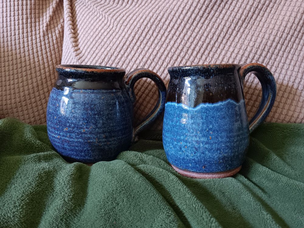 Two handmade mugs, slightly potbellied, with blue glaze covering most of the mug and black-brown glaze seeping down from the top quarter. The left mug is noticeably shorter and wider than the right, but they are clearly similar works made by the same hand. Like sisters, perhaps. 