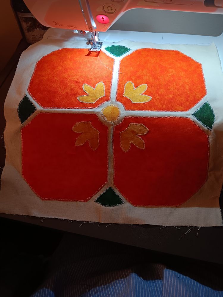 A square quilt block draped over the deck of a sewing machine. It's a flower with four orange petals, a yellow center, yellow highlights on each petal, and four green leaves peeking out around the edges. The pieces have been attached by applique.