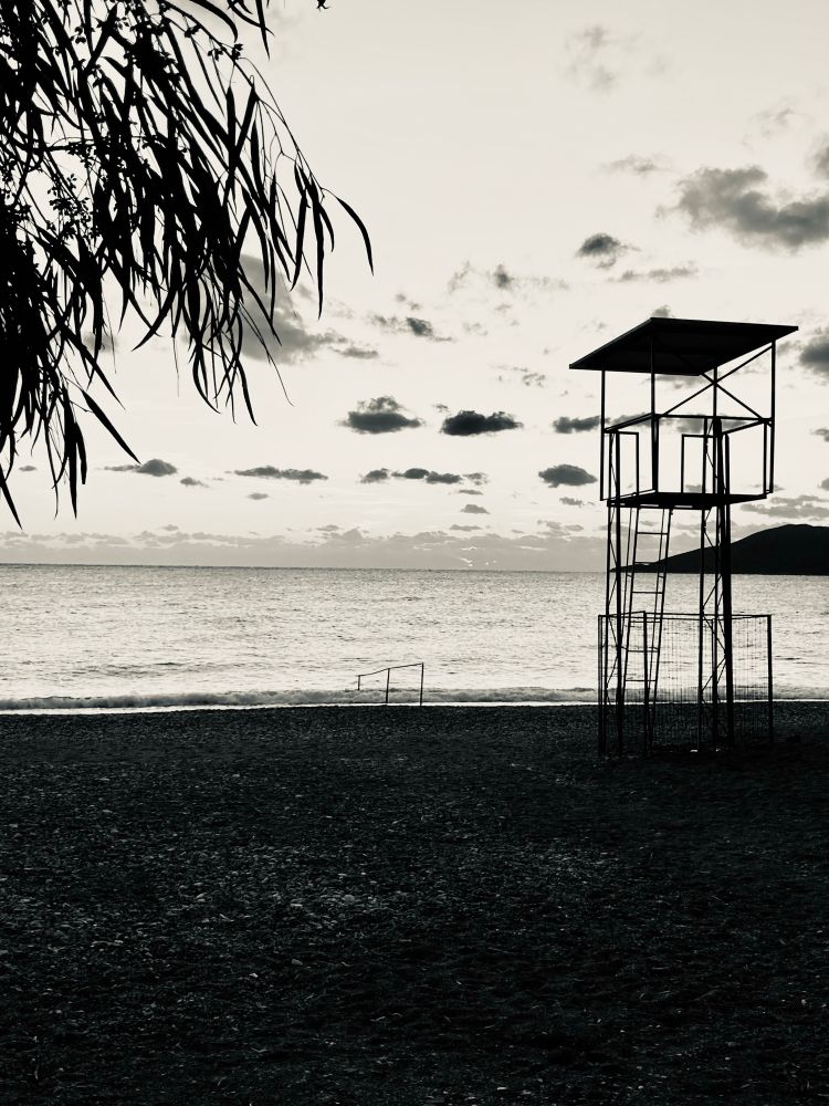 This photo was taken at dawn in Greece. It’s a black and white image of a beach shore, the flat sea in the background with bubbling clouds forming above. At the front a palm tree branch is just about in the frame together with a metal framed lifeguard lookout tower overlooking the water 