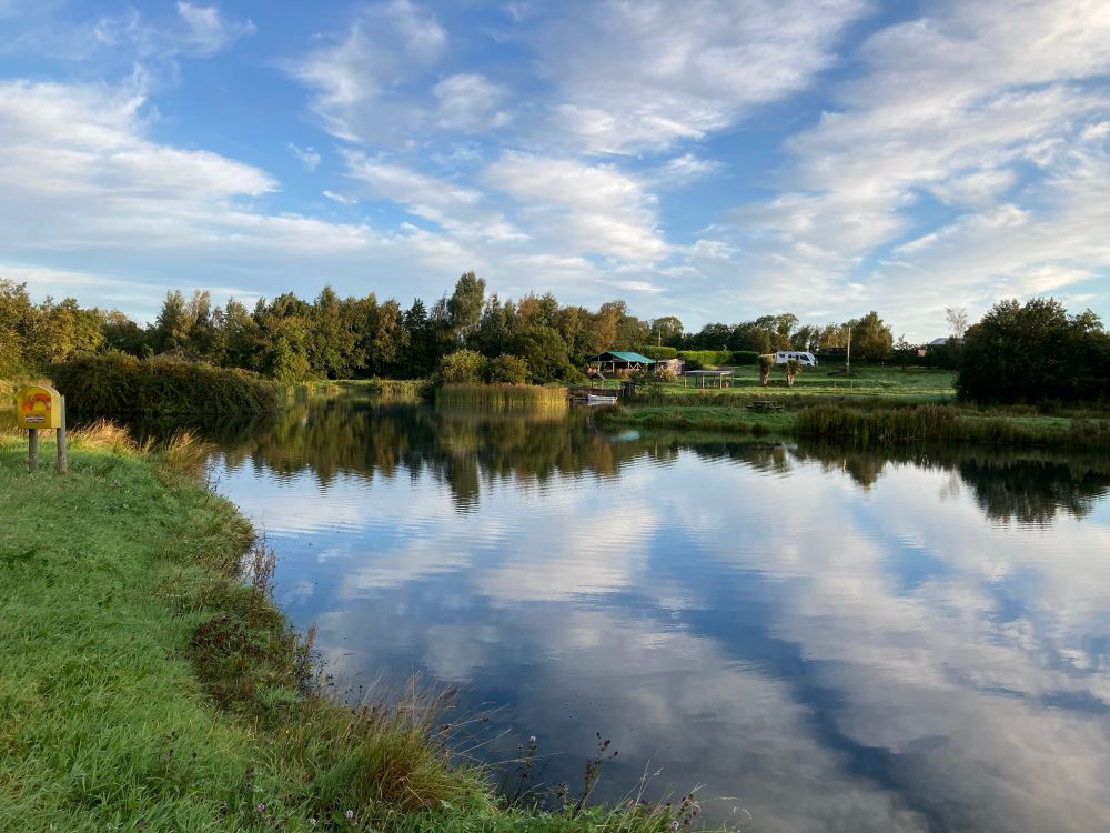 The photo of greens, whites and blues shows a lake surrounded by grass and trees. The water is still and clear and a mirror reflection of the clouds in the sky. 