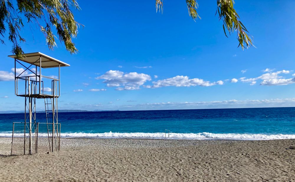 The photo was taken in Greece. A Nearly cloudless blue sky meets a two tone blue sea with small white waves lapping the golden colour sandy beach. 