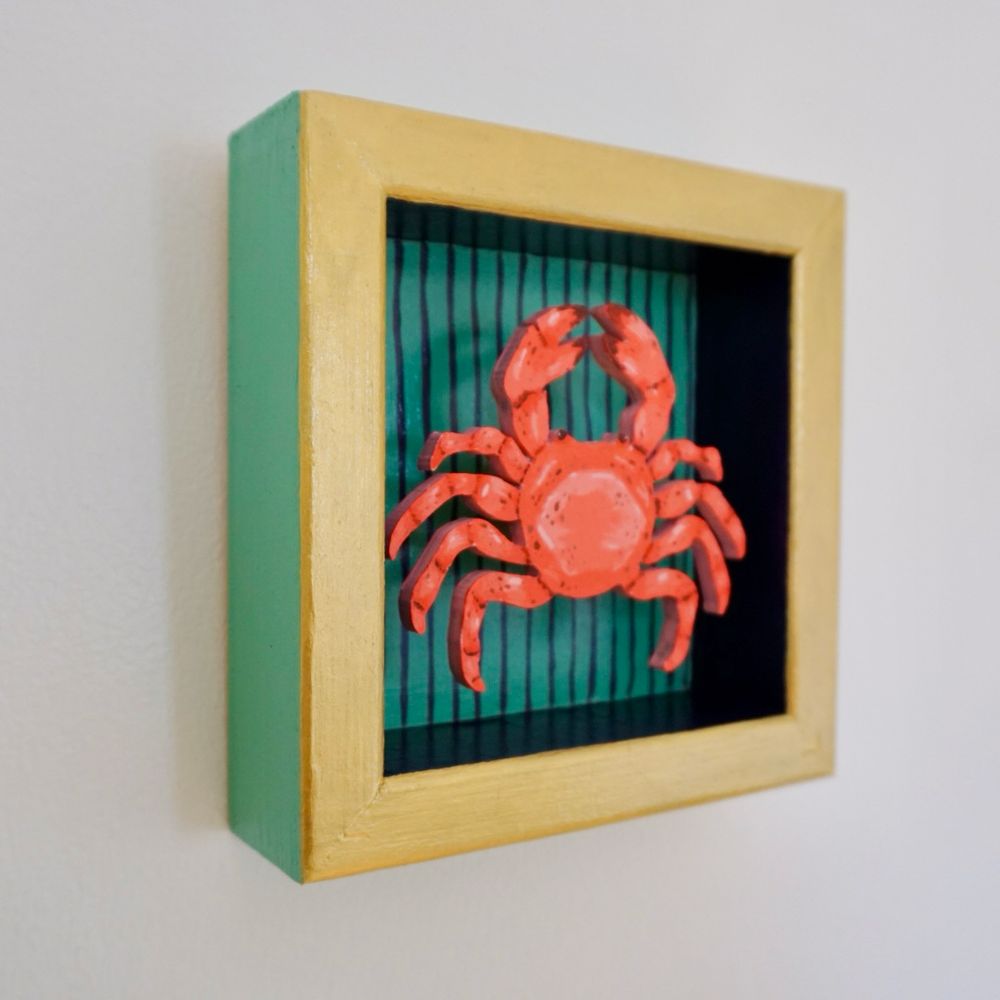 Side view of a red crab in a box that has green and dark purple stripes with a gold border 