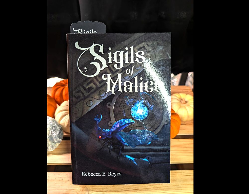 Photo of Sigils of Malice in Paperback, with a bookmark sticking out. There's pumpkins in the background.

The cover is of a magical beetle walking on magical technology and a parking crystal.