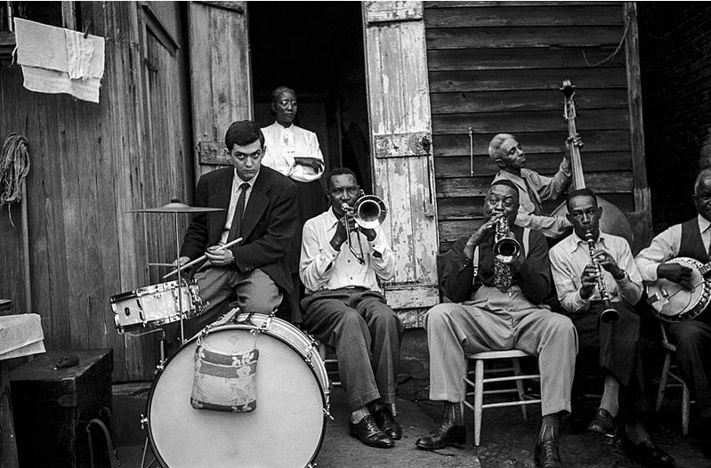 Young Stanley Kubrick, in jacket and tie, tentatively plays a snare drum next to George Lewis' band. They're posing on what appears to be the back patio of a weathered wooden house.
